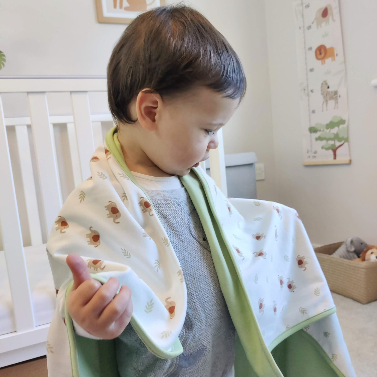 Toddler with a cotton blanket draped over his shoulders looking curious. There are safari toys and a height chart in the background