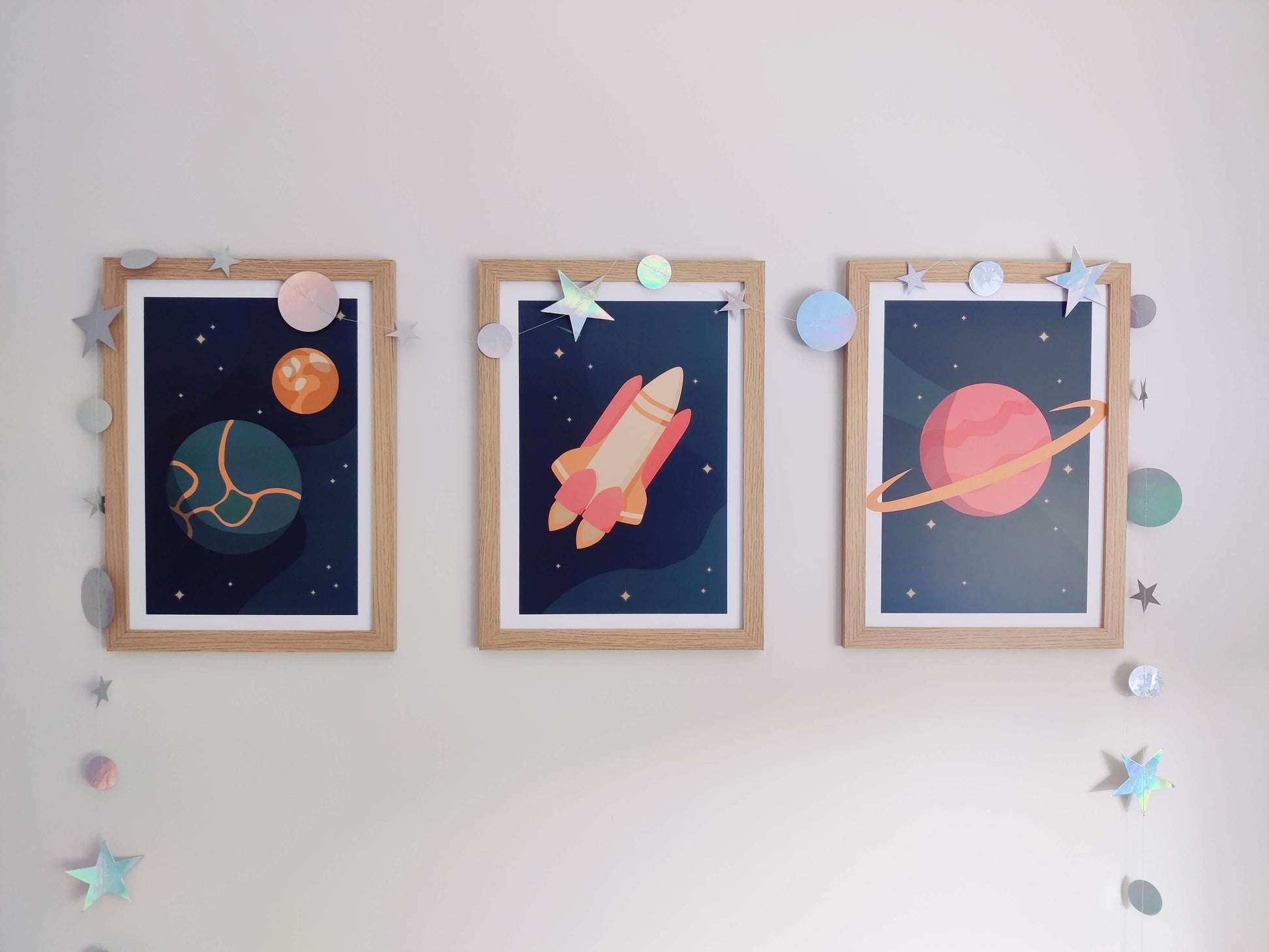 Three bold space-themed prints hanging on a wall with a iridescent star and moon garland