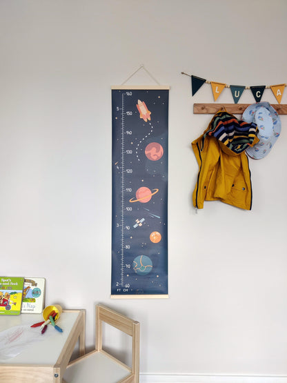 Canvas height chart with outer space theme hanging on a wall, next to coat hooks and a children’s table and chairs. There are planets, a satellite and rocket pictured on the height chart.